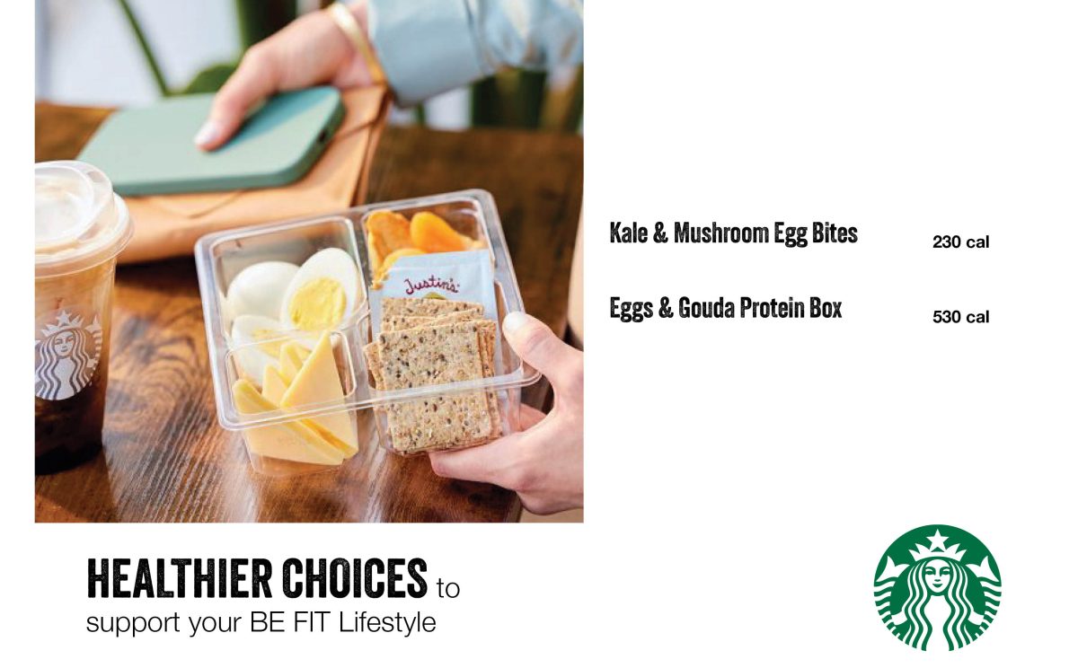 Person holding a bento-style snack box with sliced hard-boiled eggs, cheese, crackers and fruit beside an iced Starbucks drink, with menu text for Kale & Mushroom Egg Bites and Eggs & Gouda Protein Box.