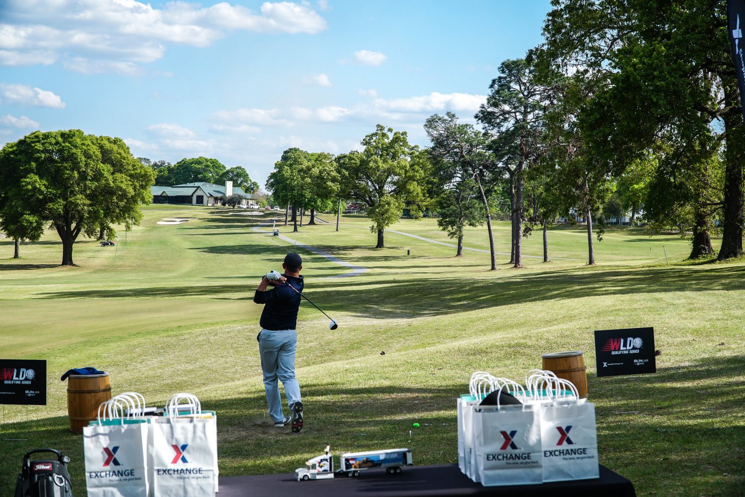 A man in a blue hat, blue shirt and white pants swings a golf club. A table with white bags labeled "Exchange" and a replica toy eighteen-wheeler sits in the foreground. 