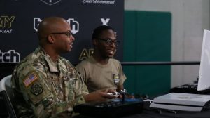 Two Soldiers play video games in front of a background that says Army Entertainment and Twitch.