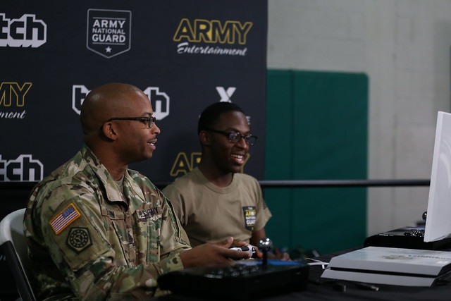 Two Soldiers play video games in front of a background that says Army Entertainment and Twitch. 