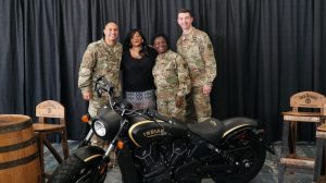 Three Soldiers in uniform stand with an Army veteran behind an Indian motorcycle